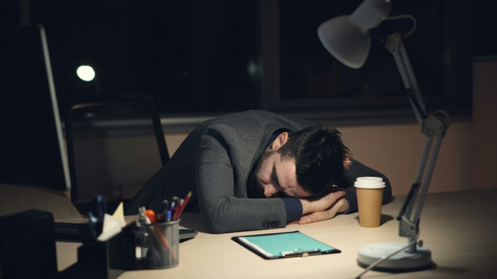 A man in a suit sleeps on his desk in a dimly lit office. A lit desk lamp, coffee cup, tablet, and pen holder are nearby, conveying exhaustion and fatigue.
